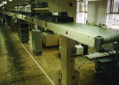 Sběrný pásový dopravník nad uličkou výrobní haly s elevátory od jednotlivých balících strojů